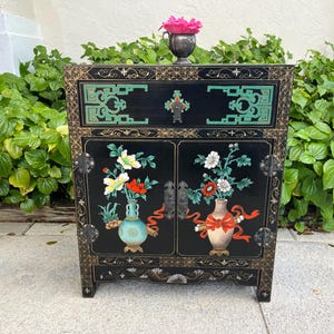 May include: Black cabinet with a drawer and two doors, featuring hand-painted floral designs in vases. The cabinet has gold trim and turquoise accents. A small silver vase with pink flowers sits on top.