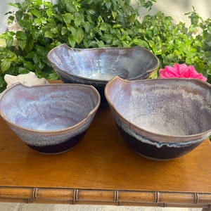 May include: Three ceramic bowls in various sizes with a speckled glaze in shades of purple, brown, and white. The bowls are sitting on a wooden table.