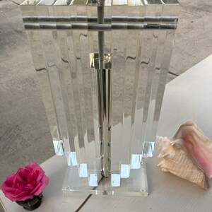 May include: A clear acrylic lamp with a silver metal pole and accents. The lamp has a rectangular top and a stepped, fan-like design. A pink rose and a conch shell are on the white surface next to the lamp.