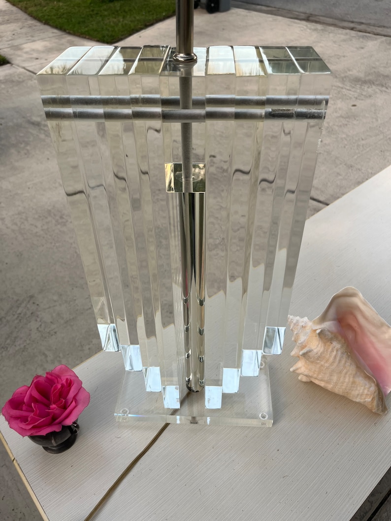 May include: A clear acrylic lamp with a silver metal pole and accents. The lamp has a rectangular top and a stepped, fan-like design. A pink rose and a conch shell are on the white surface next to the lamp.