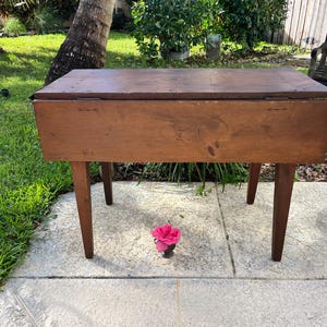 May include: A vintage wooden table with a rectangular top and four tapered legs. The table is made of a warm-toned wood and appears to be well-worn. A pink flower sits in front of the table. The table is outdoors.