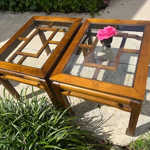 May include: A pair of rectangular side tables constructed from medium-toned wood with glass top inserts. Each table features a geometric lattice design beneath the glass surface. A small black vase containing a bright pink flower sits on one table.
