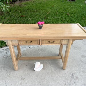 May include: A light-colored wooden desk with two drawers and a decorative top. The desk has a natural wood finish and a woven texture on the drawer fronts. A small silver vase with pink flowers sits on the desk. A large seashell rests on the ground.