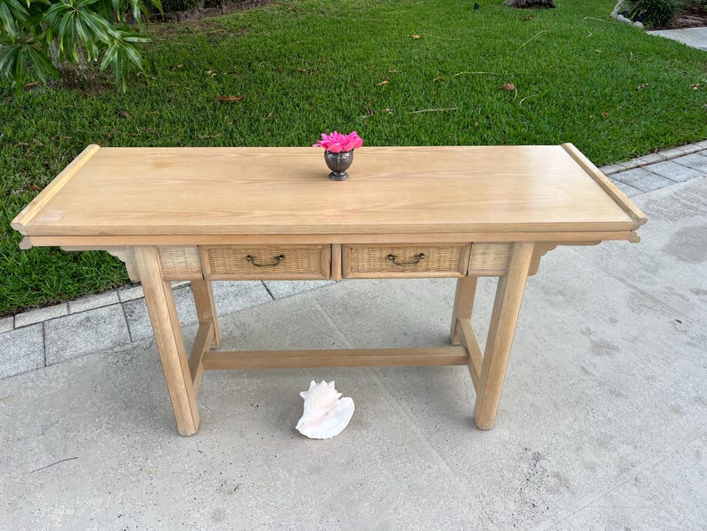 May include: A light-colored wooden desk with two drawers and a decorative top. The desk has a natural wood finish and a woven texture on the drawer fronts. A small silver vase with pink flowers sits on the desk. A large seashell rests on the ground.