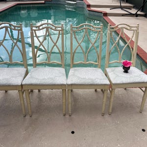 May include: Four cream-colored dining chairs with light gray patterned seats. The chairs have a bamboo-style back design. A small black vase with a pink rose sits on one chair. The chairs are arranged near a pool.