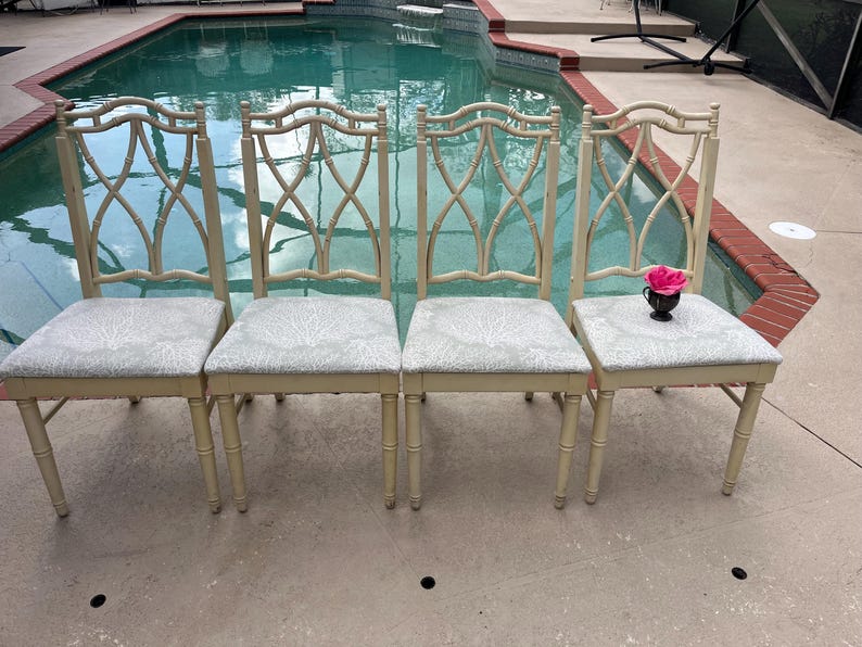 May include: Four cream-colored dining chairs with light gray patterned seats. The chairs have a bamboo-style back design. A small black vase with a pink rose sits on one chair. The chairs are arranged near a pool.