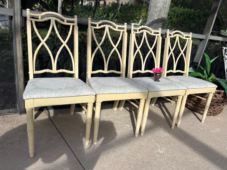 May include: Four cream-colored dining chairs with patterned gray seats. The chairs have a decorative back design and bamboo-style legs. A small silver vase with a pink flower sits on one of the seats.
