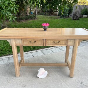 May include: A light-colored wooden desk with two drawers and woven accents. The desk has a rectangular top and angled legs. A small silver vase with pink flowers sits on top. A large seashell rests on the ground in front of the desk.