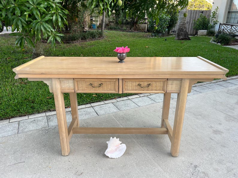May include: A light-colored wooden desk with two drawers and woven accents. The desk has a rectangular top and angled legs. A small silver vase with pink flowers sits on top. A large seashell rests on the ground in front of the desk.