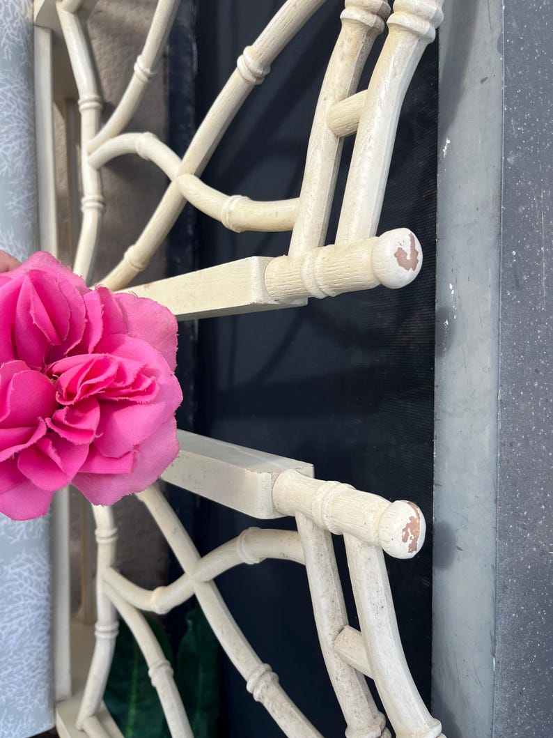 May include: Off-white, bamboo-style headboard with a lattice design. The headboard has a distressed finish, with visible wear on the rounded end pieces. A bright pink flower is in the foreground.