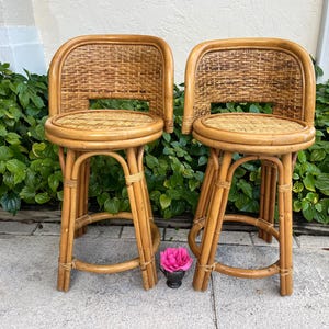May include: Two bamboo bar stools with woven backs and seats. The stools have a natural wood finish and a curved design. The stools are placed in front of green foliage. A pink flower sits between the stools.