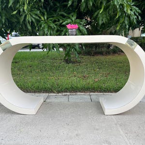 May include: A cream-colored, modern console table with a unique, curved design. The table has a rectangular top and base, with a rounded, open center. The ends of the table have gold-colored accents. A small vase with a pink flower sits on top.
