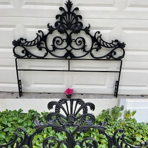 May include: Ornate black metal headboard with scrolling floral and leaf designs. The headboard has a decorative top with a fan-like element and a lower horizontal bar. A pink rose is placed below the headboard.