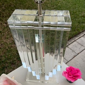 May include: A clear acrylic table lamp with a rectangular shade and a silver-toned metal base. The shade is composed of vertical, rectangular panels. A pink flower in a black pot and a seashell are next to the lamp.