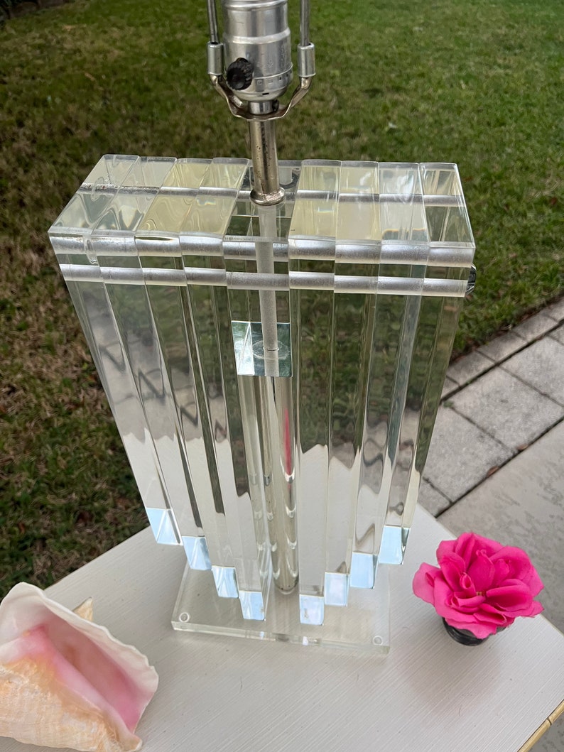 May include: A clear acrylic table lamp with a rectangular shade and a silver-toned metal base. The shade is composed of vertical, rectangular panels. A pink flower in a black pot and a seashell are next to the lamp.