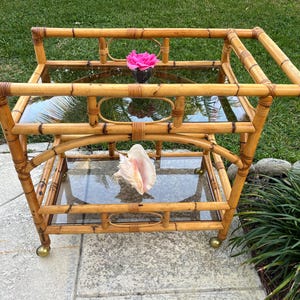 May include: A two-tiered bamboo serving cart with glass shelves and brass wheels. The cart has a pink flower in a small vase on the top shelf and a large seashell on the bottom shelf.