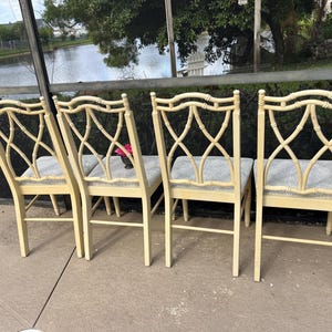 May include: Four cream-colored bamboo-style chairs with gray upholstered seats. The chairs have an open, crisscross design on the backrests. The chairs are arranged on a concrete patio, with a view of a lake and trees in the background.