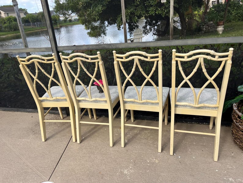 May include: Four cream-colored bamboo-style chairs with gray upholstered seats. The chairs have an open, crisscross design on the backrests. The chairs are arranged on a concrete patio, with a view of a lake and trees in the background.