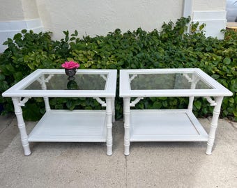 May include: Two white side tables with glass tops and lower shelves. The tables have a bamboo-style design and are placed in front of green foliage. A pink flower sits on one table. Each table is approximately 24 inches wide and 20 inches deep.
