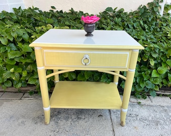 May include: A yellow side table with a white top and a drawer. The table has a lower shelf and a silver drawer pull. A small, dark vase with a pink flower sits on top. The table is 24 inches wide, 16 inches deep, and 24 inches high.