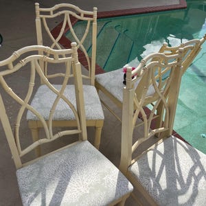 May include: Four cream-colored dining chairs with patterned seat cushions. The chairs have a bamboo-style design on the backrests and legs. The seat cushions feature a light gray and white coral pattern. The chairs are arranged near a swimming pool.