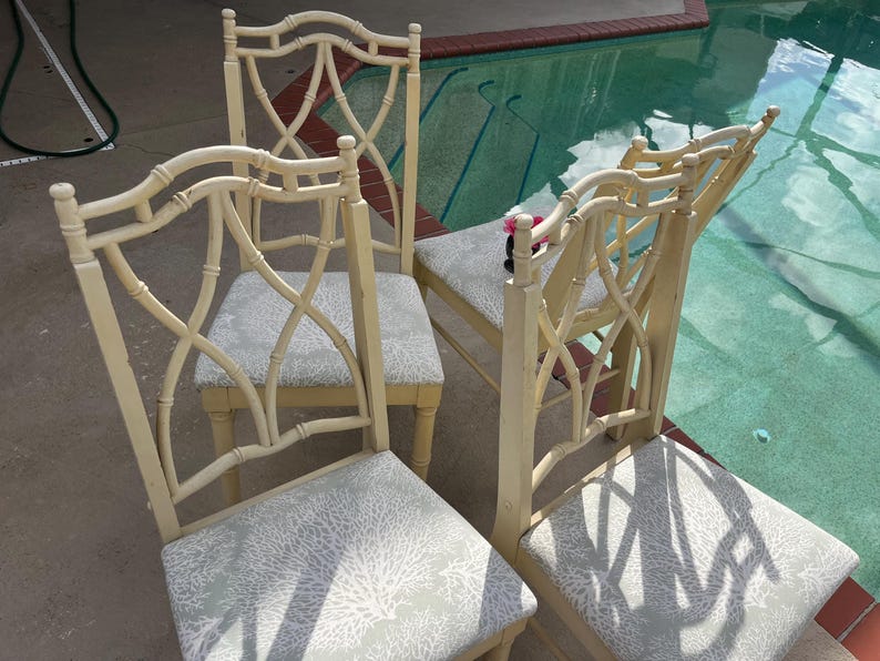 May include: Four cream-colored dining chairs with patterned seat cushions. The chairs have a bamboo-style design on the backrests and legs. The seat cushions feature a light gray and white coral pattern. The chairs are arranged near a swimming pool.