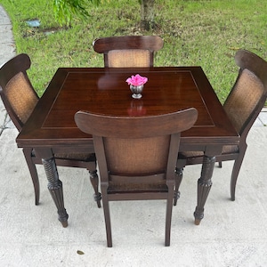 May include: A square dark brown wooden dining table with four matching chairs. Each chair features a curved backrest with a woven cane insert. The table has four carved, fluted legs and a smooth surface, topped with a small silver vase holding a pink flower.