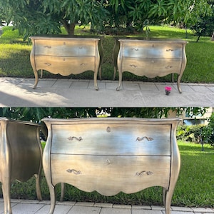 May include: Three antique-style nightstands with two drawers each. The nightstands have a gold and silver finish with ornate hardware and curved legs. The background features green foliage and a concrete surface.