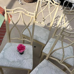 May include: Four cream-colored bamboo-style chairs with light blue seats featuring a white coral pattern. A pink rose in a small black cup sits on one chair. The chairs have an open, crisscross back design.