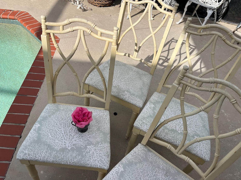 May include: Four cream-colored bamboo-style chairs with light blue seats featuring a white coral pattern. A pink rose in a small black cup sits on one chair. The chairs have an open, crisscross back design.
