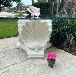 May include: A coffee table with a circular glass top supported by a white, sculpted base shaped like a large seashell. The base features intricate, radiating ridges and a textured, rope-like border along the bottom edge, resting on a flat surface.
