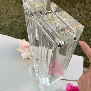 May include: A clear acrylic lamp with a rectangular prism design. The lamp features a silver-colored metal neck and a clear base. A pink flower and a seashell are next to the lamp.