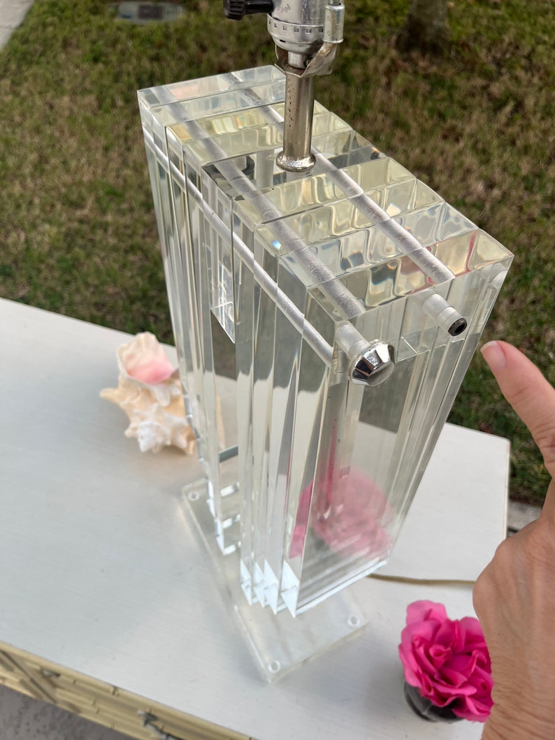 May include: A clear acrylic lamp with a rectangular prism design. The lamp features a silver-colored metal neck and a clear base. A pink flower and a seashell are next to the lamp.