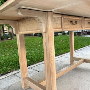 May include: A light-colored wooden table with three small drawers featuring woven rattan fronts. The table has a rectangular top and sturdy legs, showcasing a natural wood grain finish. The drawers have small, dark-colored handles.
