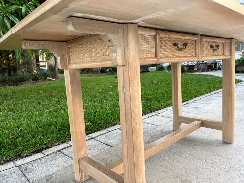 May include: A light-colored wooden table with three small drawers featuring woven rattan fronts. The table has a rectangular top and sturdy legs, showcasing a natural wood grain finish. The drawers have small, dark-colored handles.