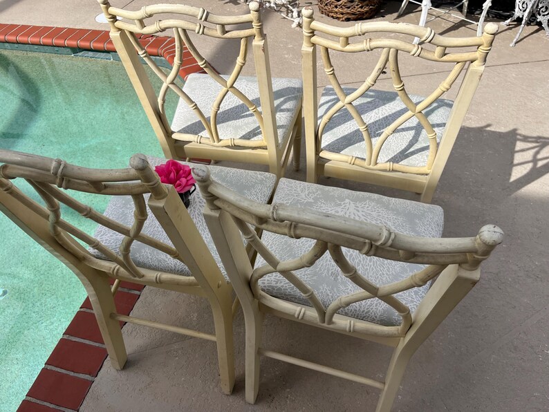 May include: Four bamboo-style chairs with light beige frames and patterned seat cushions. The chairs have a decorative back design. A pink flower sits on one chair. The chairs are outdoors near a pool.