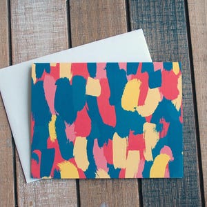 May include: A greeting card with an abstract design in teal, pink, and yellow brushstrokes. It is accompanied by a white envelope, set against a wooden background.