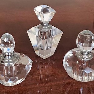 Puede incluir: Tres frascos de perfume de cristal transparente con tapones facetados y collares plateados. Los frascos tienen diferentes formas: un cuadrado alto, uno redondo y otro ovalado aplanado. Se encuentran sobre una superficie marrón oscuro.