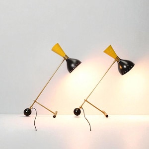 May include: Two mid-century modern desk lamps. Each lamp has a black shade with a yellow top, a brass-colored arm, and a black base. The lamps are angled towards the viewer, casting a warm glow.