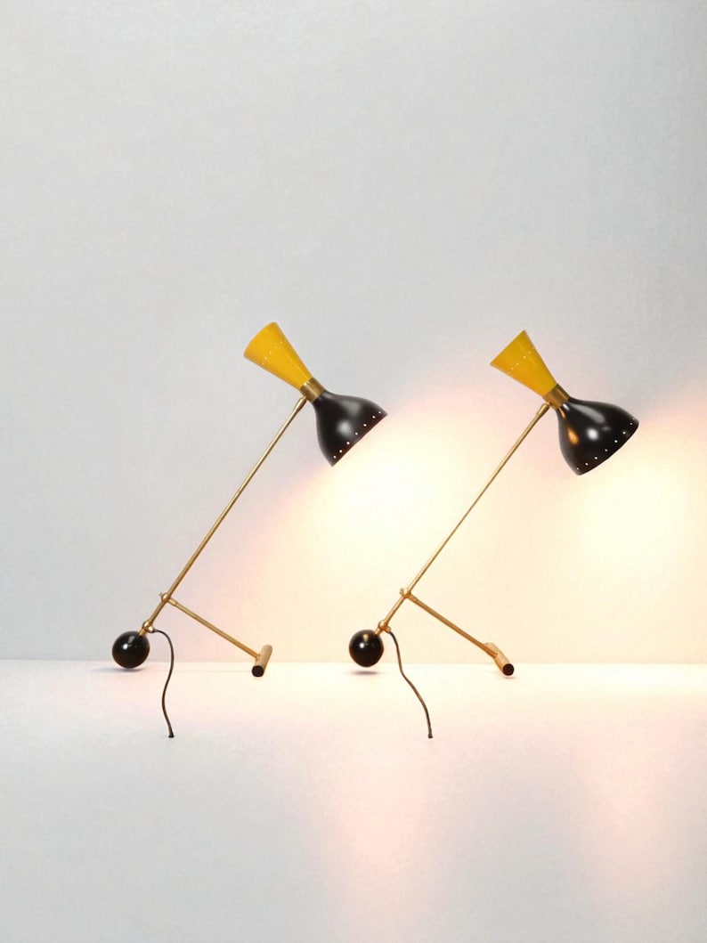 May include: Two mid-century modern desk lamps. Each lamp has a black shade with a yellow top, a brass-colored arm, and a black base. The lamps are angled towards the viewer, casting a warm glow.