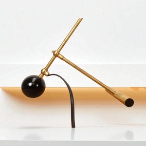 May include: A modern, minimalist desk lamp with a black spherical shade and a brass-colored, articulated arm. The lamp features a curved black support and a cylindrical end. The design is sleek and contemporary.