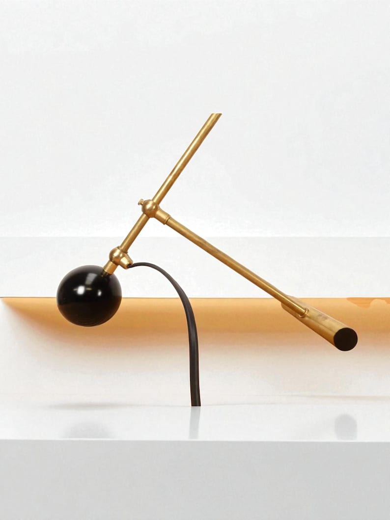 May include: A modern, minimalist desk lamp with a black spherical shade and a brass-colored, articulated arm. The lamp features a curved black support and a cylindrical end. The design is sleek and contemporary.