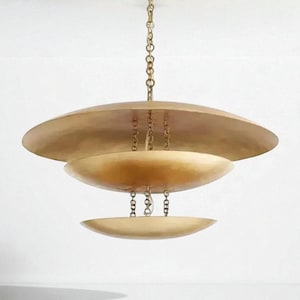 May include: A gold-toned, tiered chandelier with three circular, saucer-like shades suspended by chains. The light fixture has a mid-century modern design, with a warm, metallic finish. The shades are arranged in descending order, creating a layered effect.
