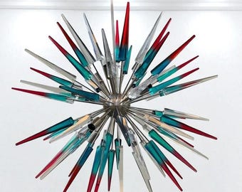 May include: A modern chandelier with a starburst design. It features numerous clear, teal, and red glass spikes radiating from a central silver sphere. The fixture is suspended from a thin, gold-colored rod.