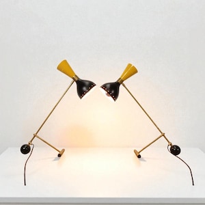 Może przedstawiać: Dwie nowoczesne lampy biurkowe z połowy wieku. Każda lampa ma czarny klosz z żółtą górą, ramię w kolorze mosiądzu i czarną kulę u podstawy. Lampy są ustawione naprzeciwko siebie, ze świecącym światłem.