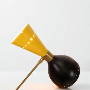 May include: A mid-century modern desk lamp with a yellow conical shade and a black, rounded base. The lamp features a brass arm and accents, and small perforations in the shade and base that emit light. The design is sleek and minimalist.