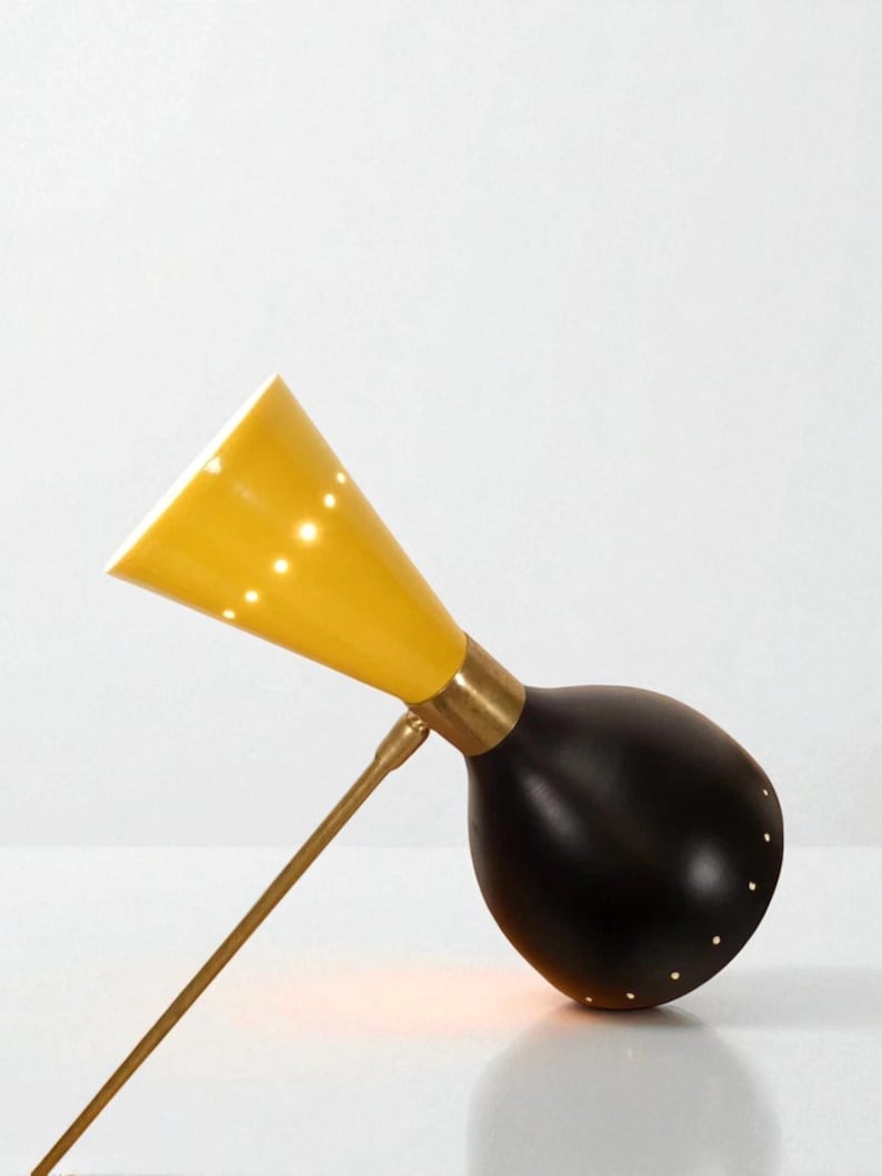 May include: A mid-century modern desk lamp with a yellow conical shade and a black, rounded base. The lamp features a brass arm and accents, and small perforations in the shade and base that emit light. The design is sleek and minimalist.