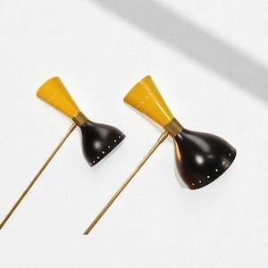 May include: Two mid-century modern wall sconces. Each sconce features a yellow conical shade and a black, bell-shaped shade. The shades are connected by a brass-colored arm. The sconces are mounted on a white wall.