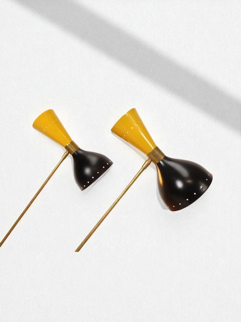 May include: Two mid-century modern wall sconces. Each sconce features a yellow conical shade and a black, bell-shaped shade. The shades are connected by a brass-colored arm. The sconces are mounted on a white wall.