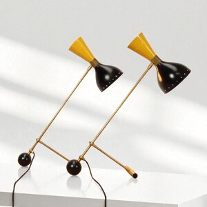 May include: Two mid-century modern desk lamps. Each lamp has a yellow conical shade, a black bell-shaped shade, and a brass-colored arm. The lamps are connected to a black base with a black sphere and a black cord.
